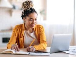 Young black woman with dreadlocks sitting at desk with laptop open and writing on pen and paper 